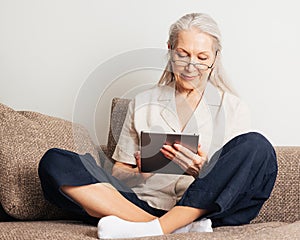 Senior woman with grey hair wearing glasses sitting with crossed legs on a couch using a digital tablet