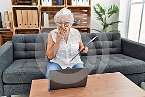 Senior woman with grey hair working at consultation office doing online therapy covering mouth with hand, shocked and afraid for