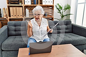 Senior woman with grey hair working at consultation office doing online therapy pointing thumb up to the side smiling happy with