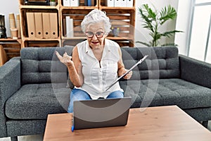 Senior woman with grey hair working at consultation office doing online therapy smiling happy pointing with hand and finger to the