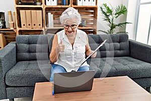 Senior woman with grey hair working at consultation office doing online therapy smiling happy and positive, thumb up doing