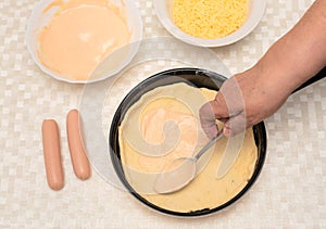 Senior woman hand with spoon adding sauce on the raw dough on black pan for homemade pizza. Other ingredients are on the
