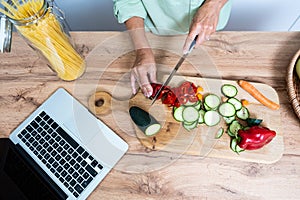 Senior woman hands preparing food in domestic kitchen view from above chopping the vegetables while using laptop computer for