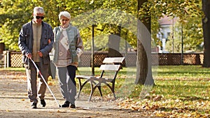 Senior woman helping her disabled blind husband in sunglasses to walk through the park