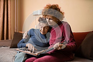 Senior woman and her adult daughter looking at photo album together on couch in living room, talking joyful discussing memories.