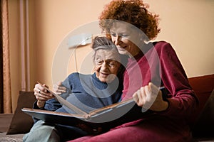 Senior woman and her adult daughter looking at photo album together on couch in living room, talking joyful discussing memories.