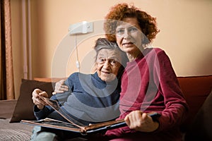 Senior woman and her adult daughter looking at photo album together on couch in living room, talking joyful discussing memories.