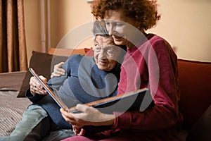 Senior woman and her adult daughter looking at photo album together on couch in living room, talking joyful discussing memories.
