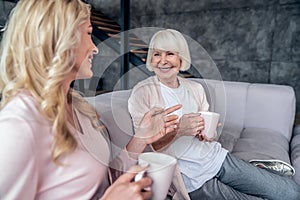 Senior woman and her attractive daughter spending time together at home. Sitting on sofa and drinking tea together. Happy Mothers