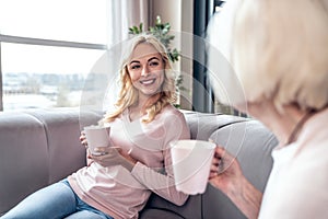 Senior woman and her attractive daughter spending time together at home. Sitting on sofa and drinking tea together. Happy Mothers