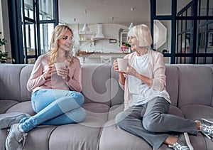 Senior woman and her attractive daughter spending time together at home. Sitting on sofa and drinking tea together. Happy Mothers