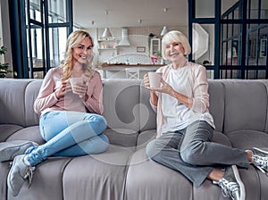 Senior woman and her attractive daughter spending time together at home. Sitting on sofa and drinking tea together. Happy Mothers