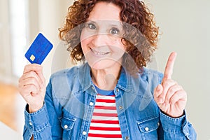 Senior woman holding credit card as payment surprised with an idea or question pointing finger with happy face, number one