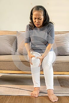 Senior woman holding her knee in pain while sitting on the sofa Profile of an unhappy old woman.