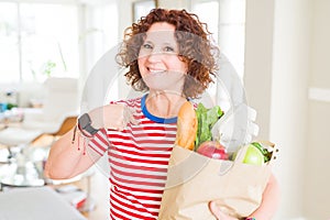 Senior woman holding paper bag full of fresh groceries from the supermarket with surprise face pointing finger to himself