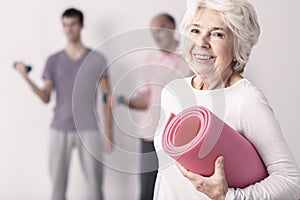 Senior woman holding pink joga mat, men exercising in background