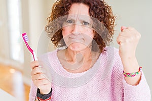 Senior woman holding pink toothbrush at dental clinic annoyed and frustrated shouting with anger, crazy and yelling with raised