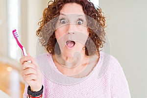 Senior woman holding pink toothbrush at dental clinic scared in shock with a surprise face, afraid and excited with fear