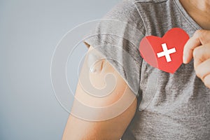 Senior woman holding red heart shape with plus sign and showing her arm with bandage after got vaccinated or inoculation