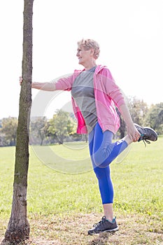 Senior woman holding tree trunk while doing leg exercise in park