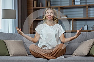 Senior woman at home resting meditating with eyes closed sitting on sofa in living room, housewife mature blonde in