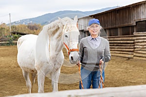 Senior woman horse breeder leading white horse through horse barn