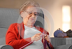 Senior woman knitting warm scarf in armchair at home