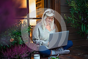 Senior woman with laptop sitting outdoors on terrace, working in the evening.