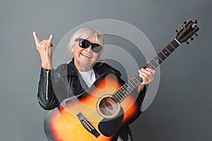 Senior woman in leather jacket and sunglasses studio standing on gray with guitar showing horns sign playful