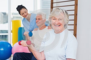 Senior woman lifting dumbbells while sitting with man and instructor