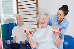 Senior woman lifting dumbbells while sitting with man and trainer