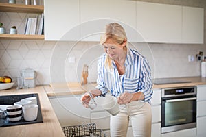 Senior woman loading or unloading dishwasher indoors at home.