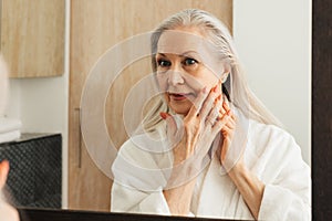 Senior woman with long grey hair touching her cheek with fingers in bathroom