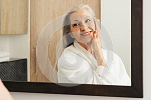 Senior woman with long grey hair touching her face with hand and looking at mirror