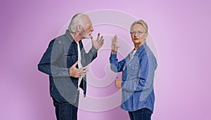 Senior woman looking at camera and showing stop gesture to furious husband arguing with her while standing isolated on purple