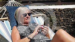 Senior woman looking on cell phone relaxing on a hammock in home garden