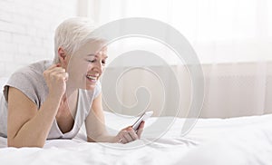 Senior woman lying on bed reading text message on phone
