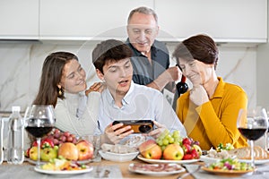 Senior woman and man celebrating family holiday with their son and his young wife at kitchen table, looking at photos on