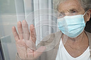 Senior woman in mask looking out of window at nursing home, view from outside