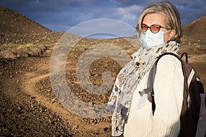 Senior woman with medical face mask walks in mountain footpath in arid landscape. Partial conquest of freedom after coronavirus