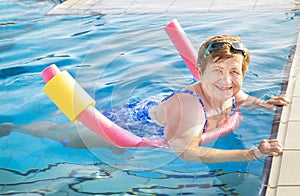 Senior woman (over age of 50) in sport goggles, swimsuit doing aqua fitness with swim noodles in swimming pool.