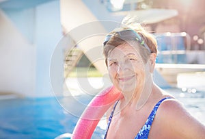 Senior woman (over age of 50) in sport goggles, swimsuit and with swim noodles near swimming pool.