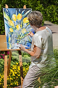 Senior woman painting flowers on canvas in garden during sunny day.