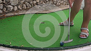 Senior woman playing minigolf, trying to put blue ball into hole, but misses many times. Detail only on her feet in sandals and