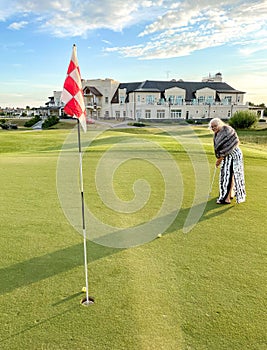 Senior woman plays golf on golf course. Background of luxury mansion. Scores ball in hole with flag