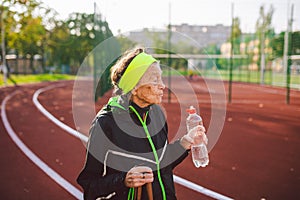 Senior woman practicing nordic walking at city stadium and stopped to quench thirst, drink water from bottle. Age, maturity,