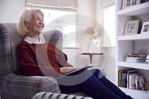 Senior Woman Reading Book Keeping Warm By Portable Radiator In Cost Of Living Energy Crisis