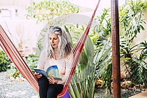 Senior woman reading book sitting on hammock at front or back yard. Elderly woman relaxing at garden. Old woman sitting on swing