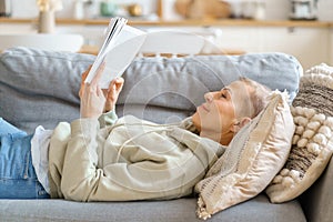 Senior woman reading book and smiling while relaxing on sofa at home