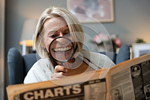 Senior woman reading newspaper with magnifying glass, unaware making funny facial expressions. Middle age retired female having